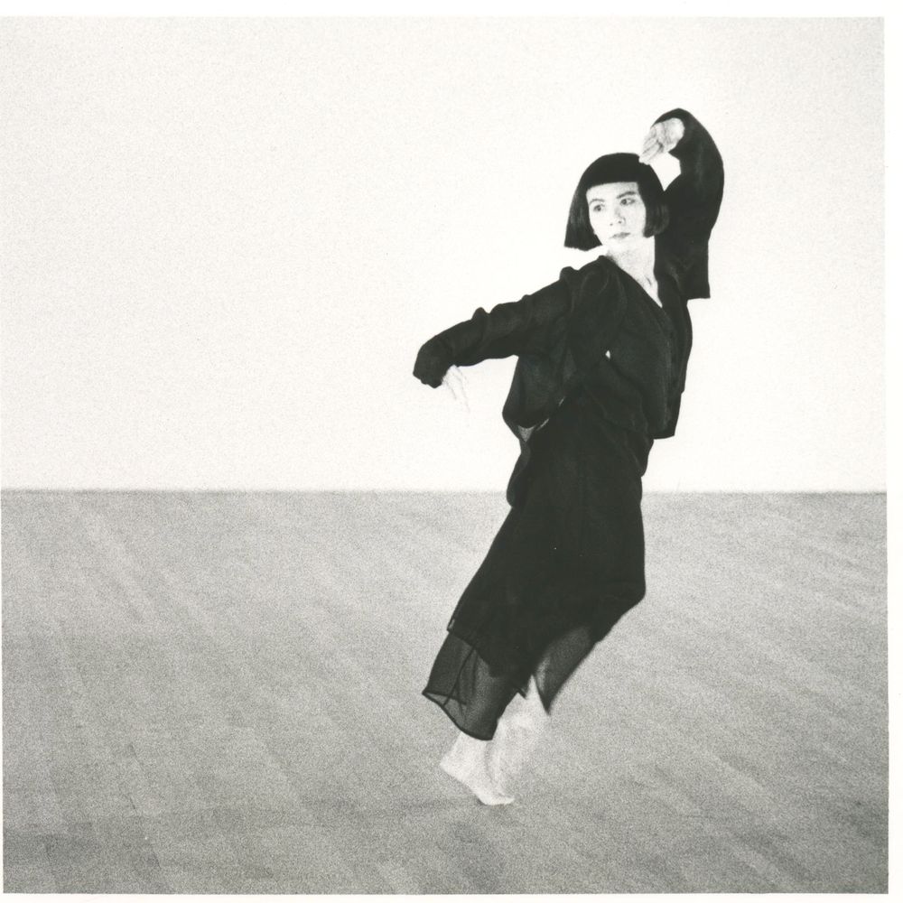 Een danseres in een expressieve pose, gekleed in een donkere, vloeiende outfit, beweegt elegant over een houten vloer in een minimalistische ruimte. De zwart-witfoto benadrukt de kracht en gratie van haar beweging.