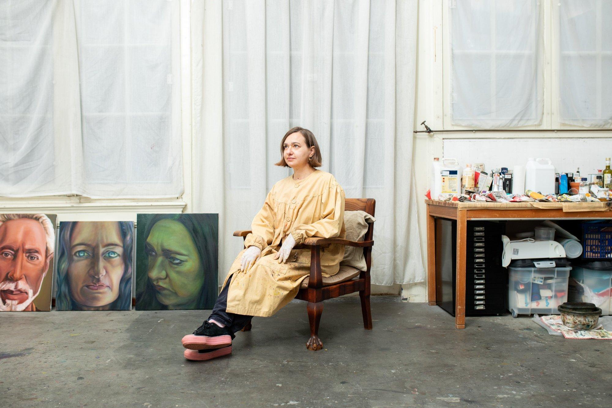 Portret van kunstenaar Ina van Zyl zittend in haar atelier met schilderijen en werktafel op de achtergrond.