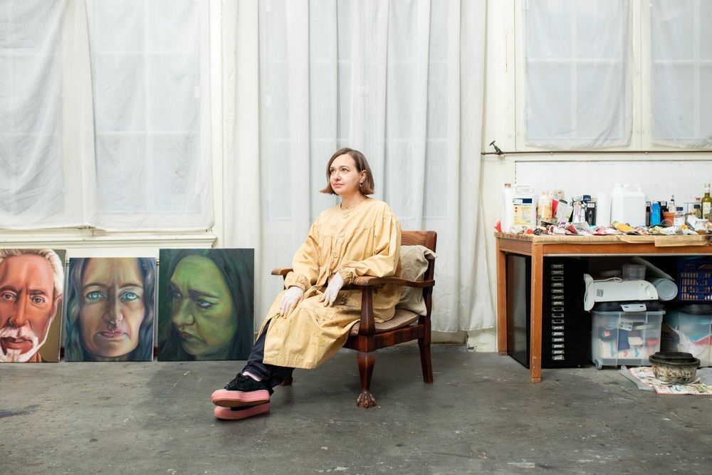 Portret van kunstenaar Ina van Zyl zittend in haar atelier met schilderijen en werktafel op de achtergrond.