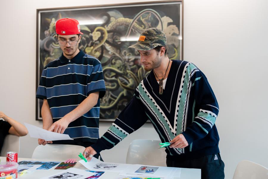 Twee deelnemers van Life Cipher bespreken hun selectie aan een tafel vol kunstafbeeldingen, met op de achtergrond een kunstwerk uit de collectie. Foto: Miko Brepols.