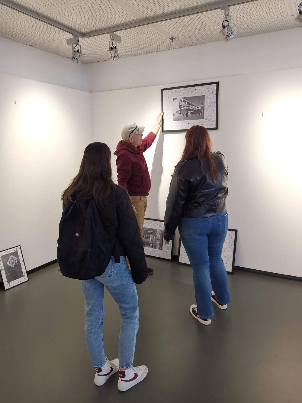 Man in hoodie hangt een ingelijste zwart-witfoto aan een witte muur terwijl twee jonge vrouwen toekijken; op de vloer staan nog meer ingelijste werken klaar voor ophanging.