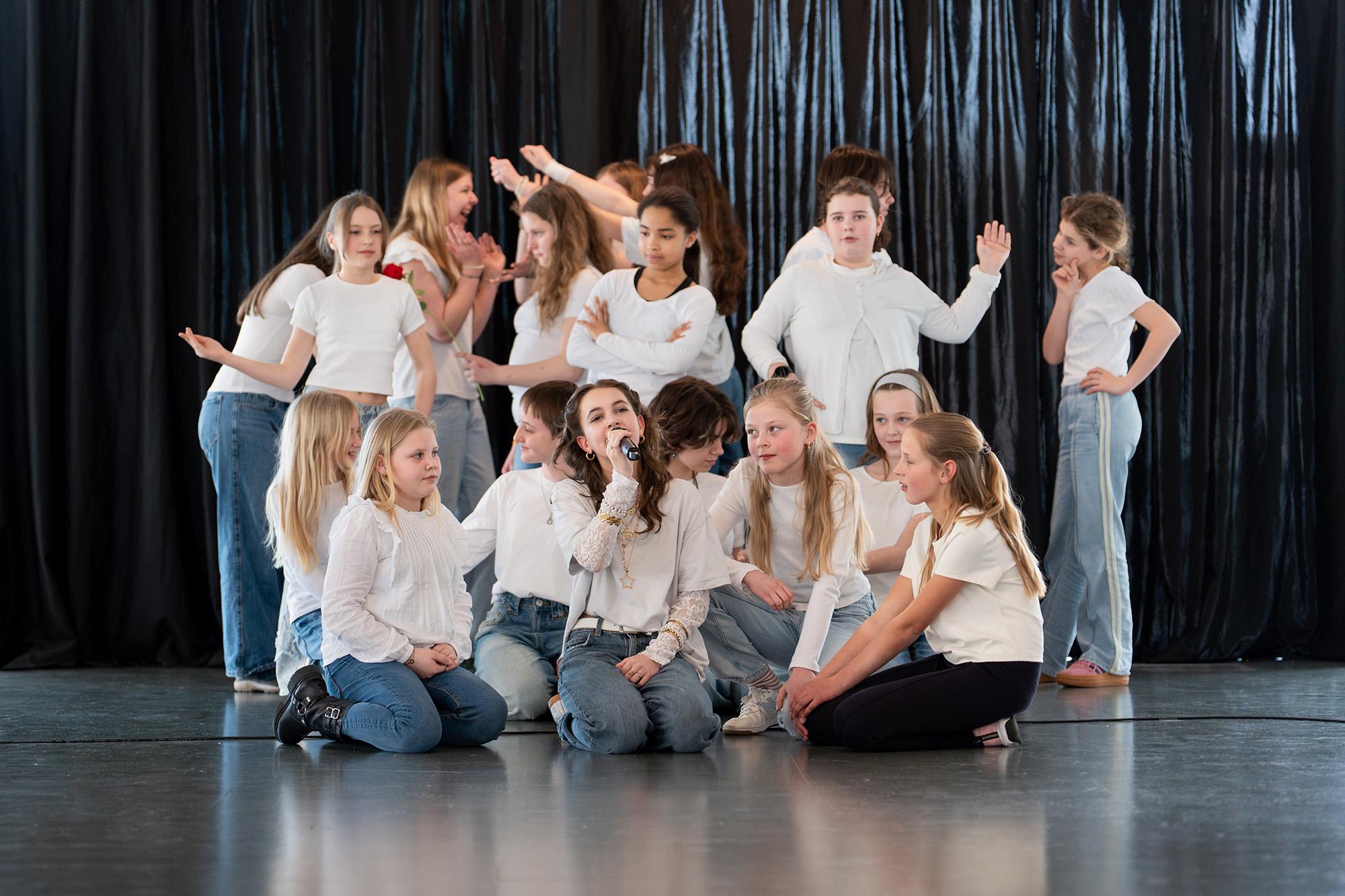 Groep kinderen in witte shirts en spijkerbroeken voert een dans- of theatervoorstelling uit voor een zwarte backdrop. Enkele kinderen zitten vooraan op de grond, terwijl anderen staand op de achtergrond poseren of bewegen.
