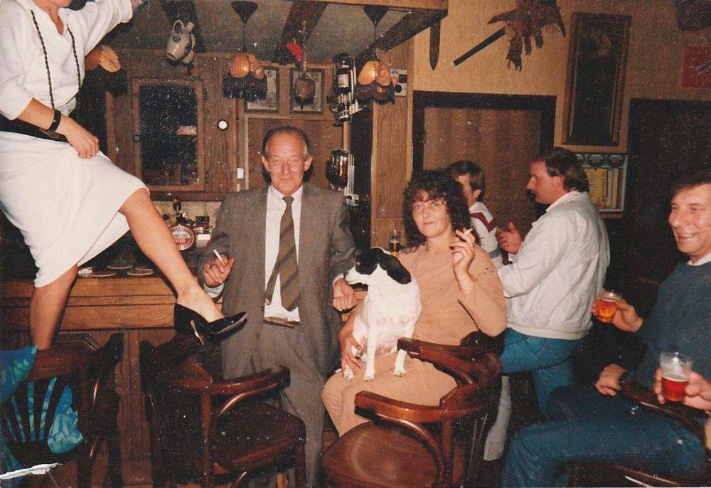 Groep mensen zit in een bruin café; een vrouw staat op een barkruk, anderen lachen, drinken en roken, met een hond op schoot bij een van de aanwezigen.