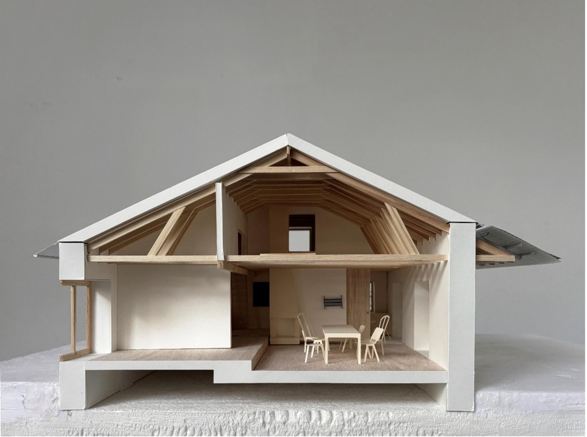 Architectuurmaquette van een woning in doorsnede, waarbij de houten dakconstructie en het interieur met tafel en stoelen zichtbaar zijn.