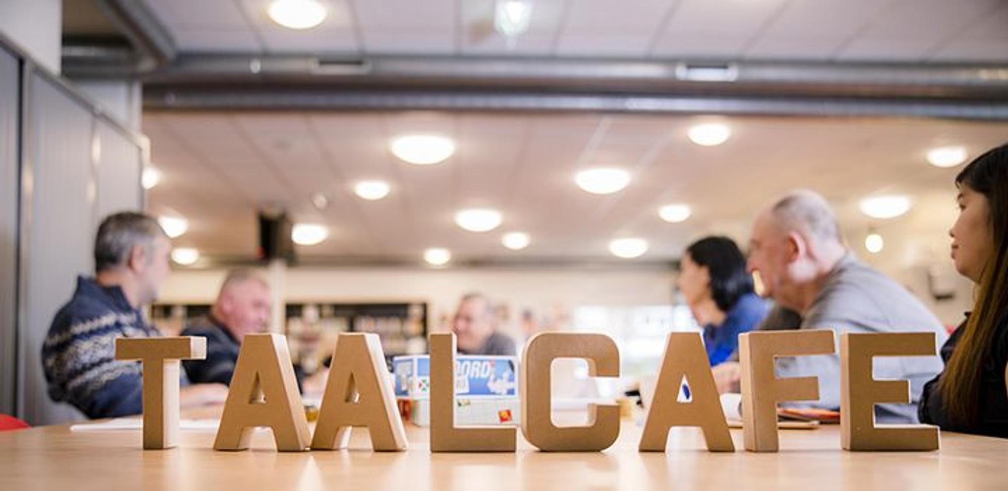 Kartonnen letters op tafel die: "Taalcafé" spellen. Op de achtergrond zitten mensen aan tafel. 