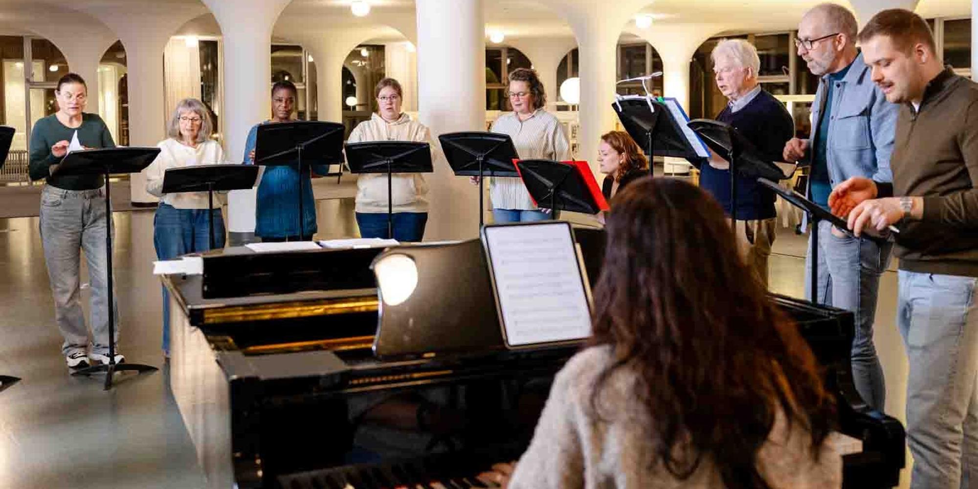 Groep zangers van SCHUNCK Popkoor Incomplete staat in een halve cirkel rond een piano terwijl iedereen uit bladmuziek zingt in een lichte ruimte met pilaren. Op de voorgrond zit een pianist bij de vleugel en op de achtergrond zijn muziekstandaards en een open interieur met ramen zichtbaar.
