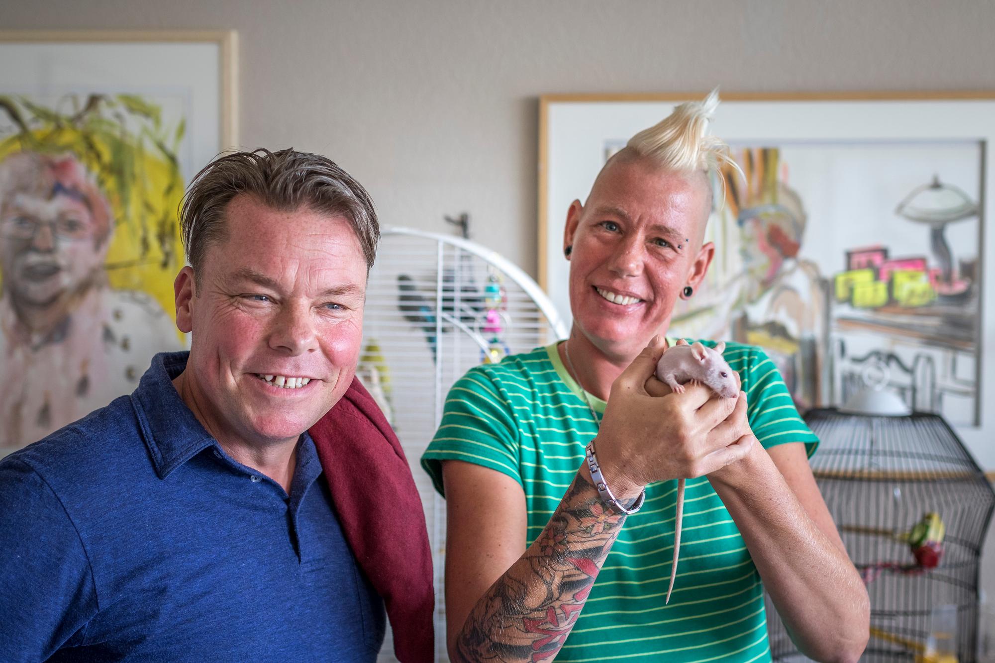 David Bade samen met een vrouw die een naakt model in haar handen vasthoudt. Op de achtergrond hangen schilderijen van David Bade aan de muur en staan twee kooien met vogels.