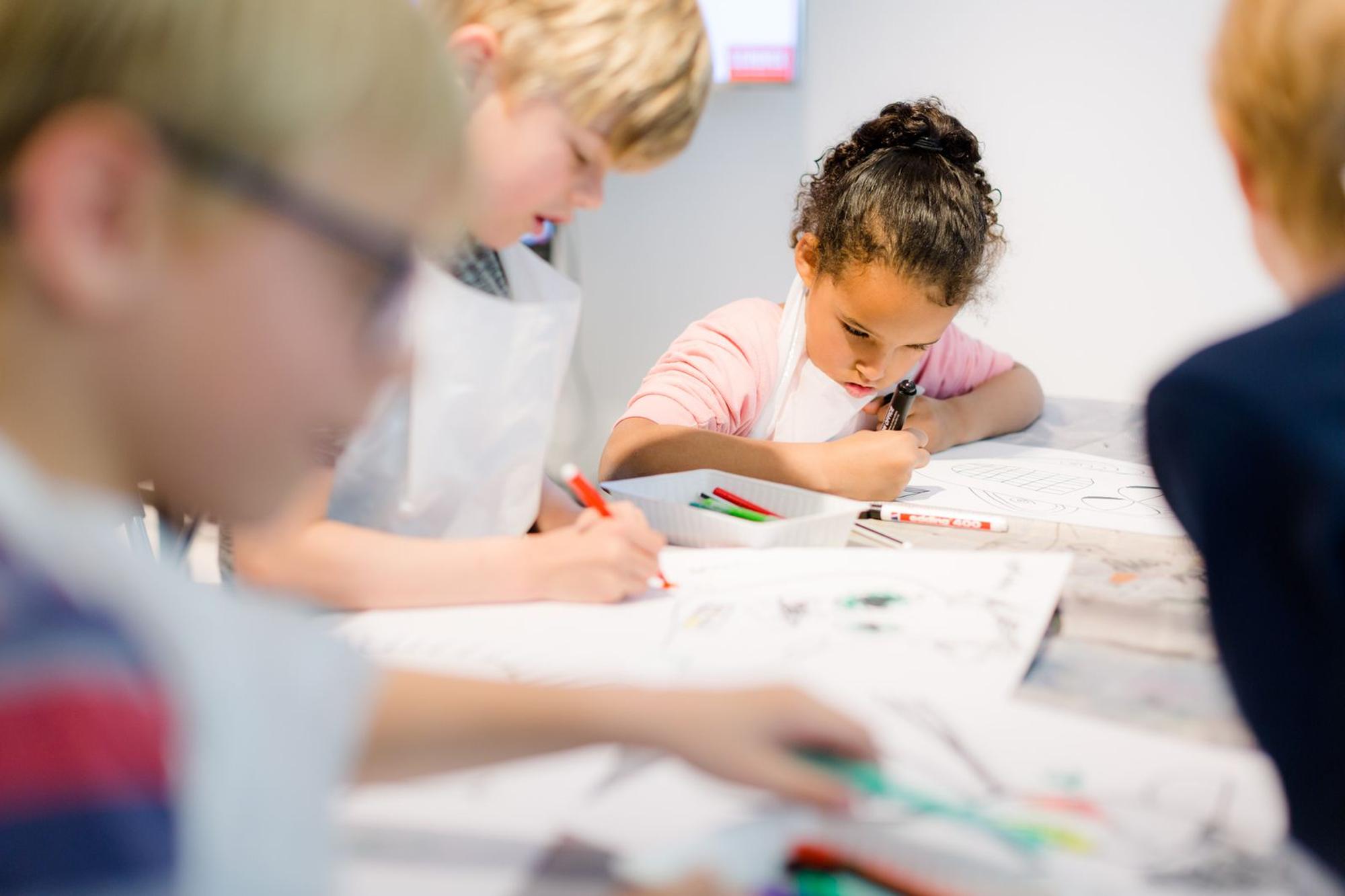 Kinderen zitten aan een tafel en kleuren geconcentreerd tekeningen in met stiften tijdens een creatieve workshop bij SCHUNCK.