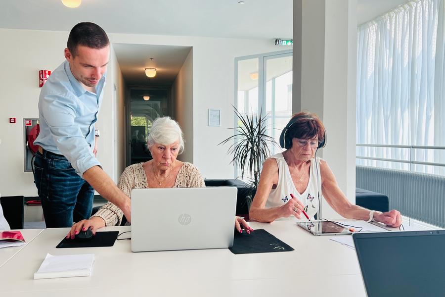 Een jonge man helpt twee oudere vrouwen tijdens een computercursus; één werkt op een laptop, de ander gebruikt een tablet met koptelefoon op.
