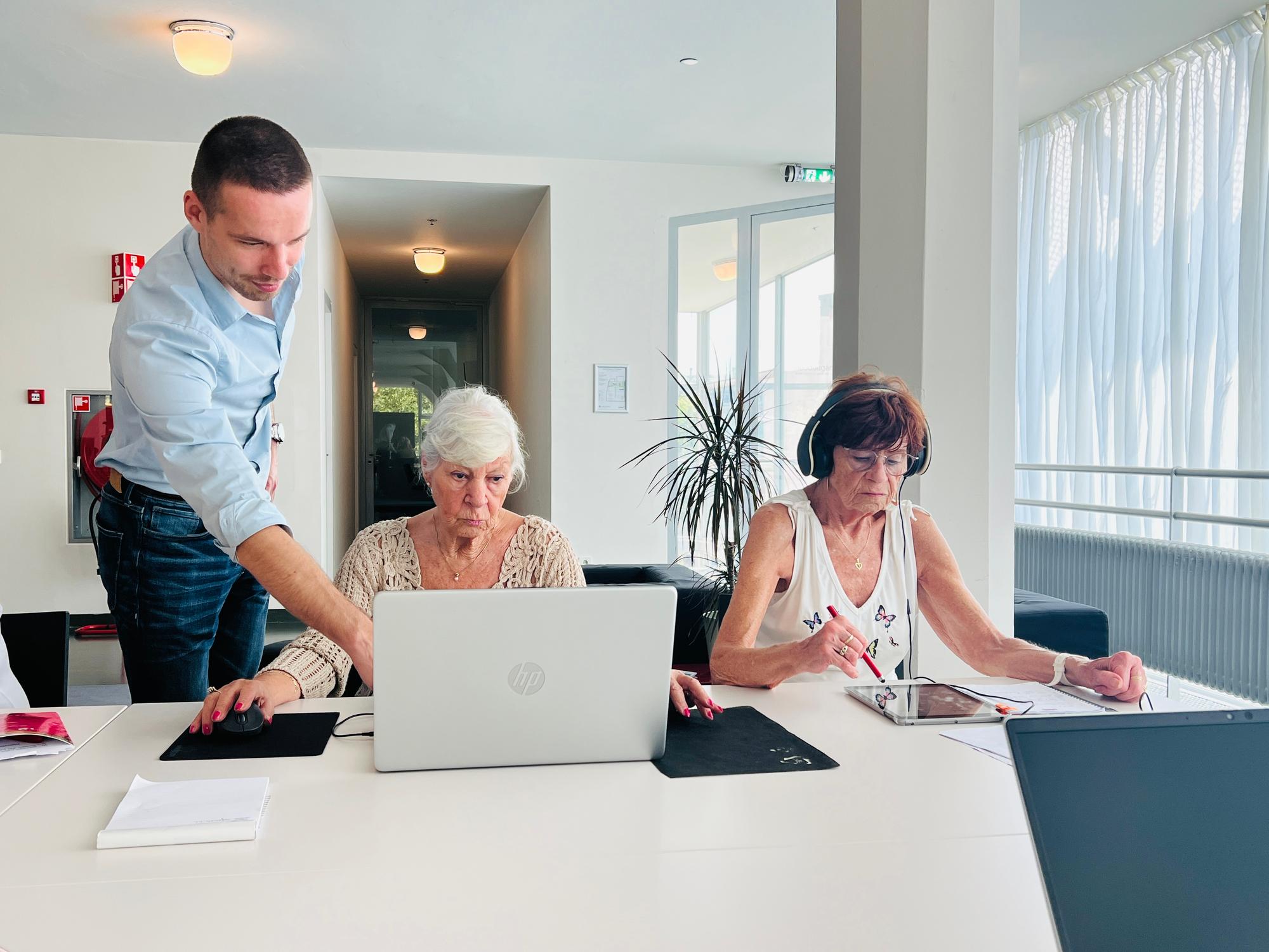 Een jonge man helpt twee oudere vrouwen tijdens een computercursus; één werkt op een laptop, de ander gebruikt een tablet met koptelefoon op.

