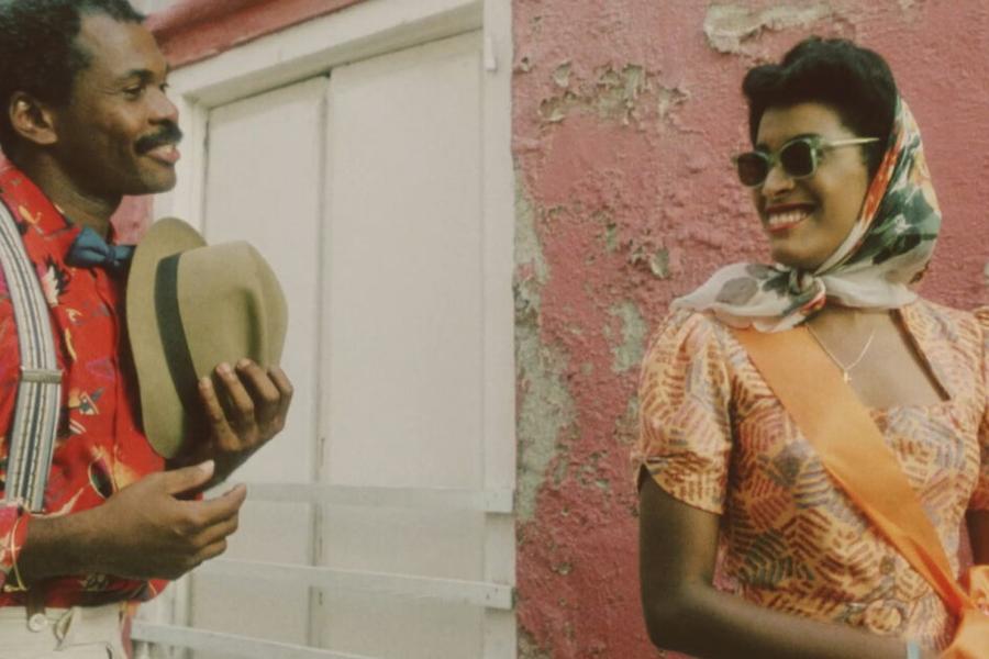 Twee personages in kleurrijke jaren ’40-outfits lopen door een zonnige straat op Curaçao, met opvallende kostuums en expressieve blikken. Still uit de film Ava & Gabriel (1990).