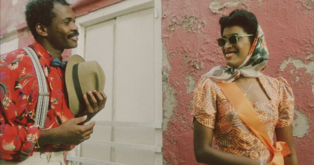 Twee personages in kleurrijke jaren ’40-outfits lopen door een zonnige straat op Curaçao, met opvallende kostuums en expressieve blikken. Still uit de film Ava & Gabriel (1990).