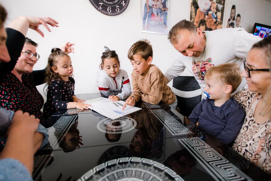 Jonge kinderen zitten met ouders en vrijwilligers aan tafel terwijl ze kijken en wijzen naar een boekje. 