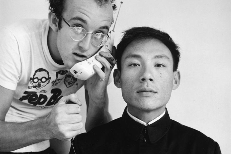 keith Haring en Tseng Kwong Chi poseren voor een witte achtergrond; links houdt een man met bril speels een grote ouderwetse mobiele telefoon tegen het hoofd van de andere man, die recht vooruit kijkt met een neutrale blik.