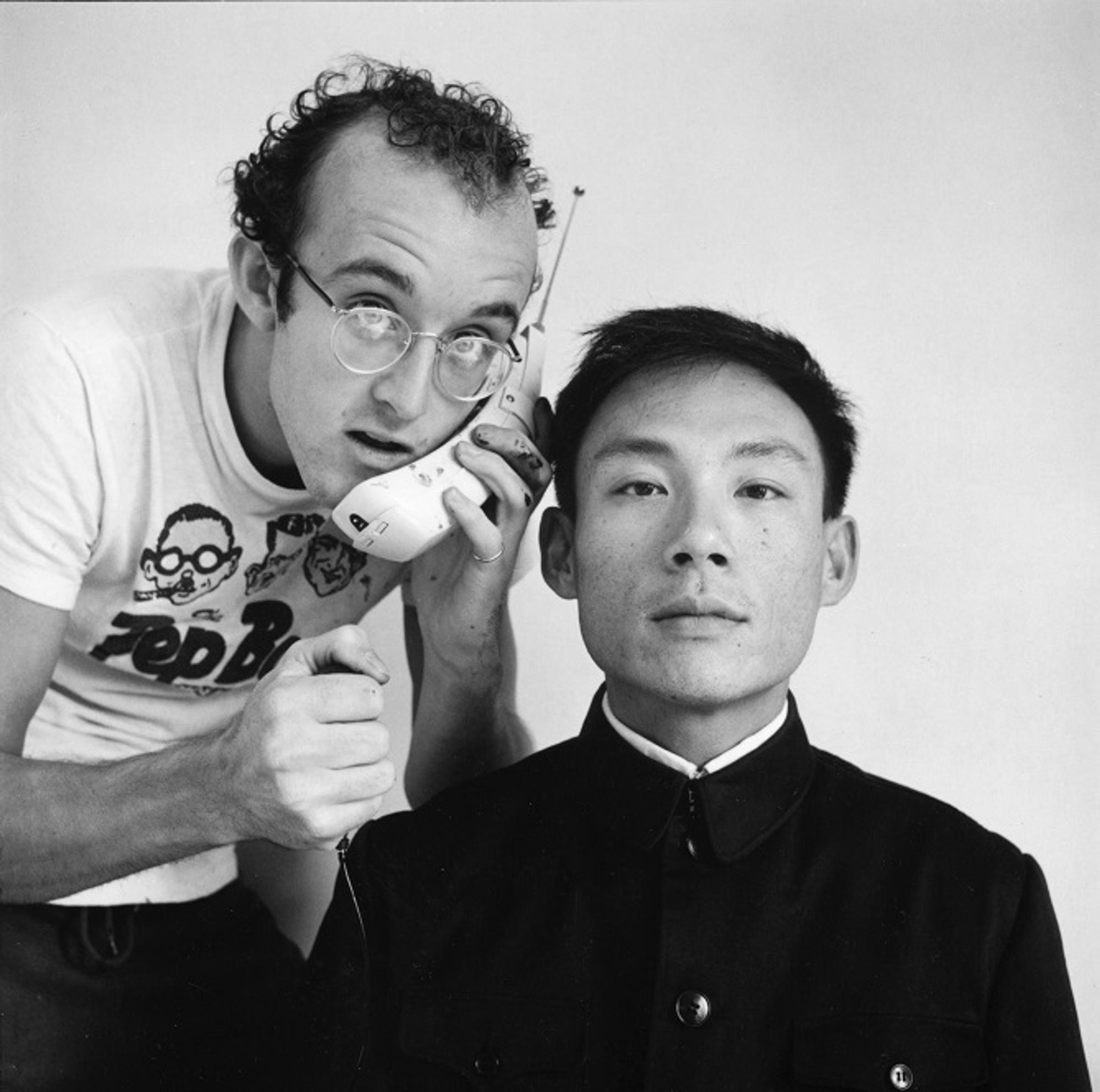 keith Haring en Tseng Kwong Chi poseren voor een witte achtergrond; links houdt een man met bril speels een grote ouderwetse mobiele telefoon tegen het hoofd van de andere man, die recht vooruit kijkt met een neutrale blik.