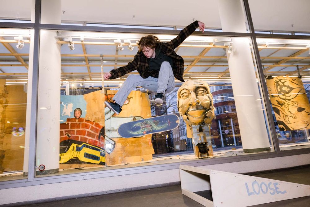 Jonge man vliegt in de lucht met skateboard, omdat ie net over een schans ging, voor de vitrine van SCHUNCK, waar kartonnen kunst te zien is. 