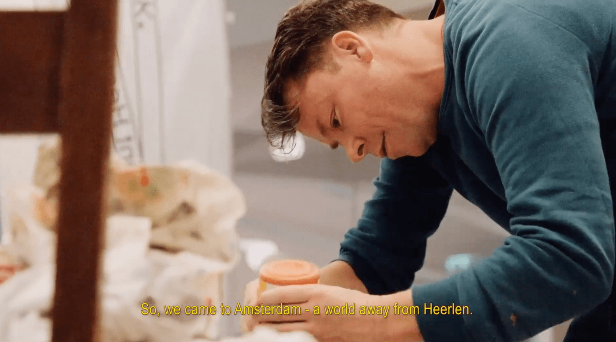 Een man in een blauwe trui buigt zich geconcentreerd over zijn werktafel, waar hij met beide handen een voorwerp vasthoudt. Op de achtergrond zijn vage materialen zichtbaar. Onderaan in beeld staat de Engelse ondertiteling: “So, we came to Amsterdam – a world away from Heerlen.”