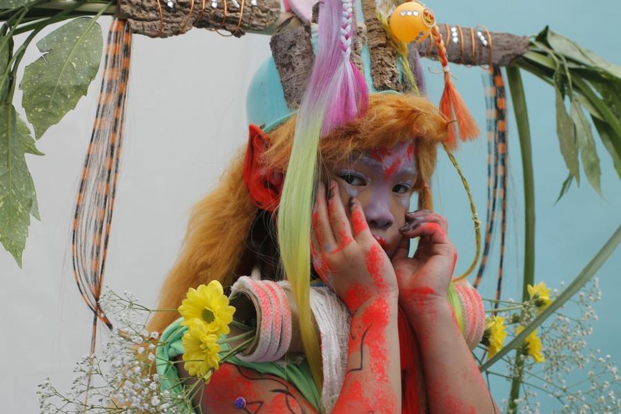 Portret van een figuur in fantasierijke kostuums met bloemen, felgekleurde verf en natuurlijke materialen, gefotografeerd tegen een lichtblauwe achtergrond.
