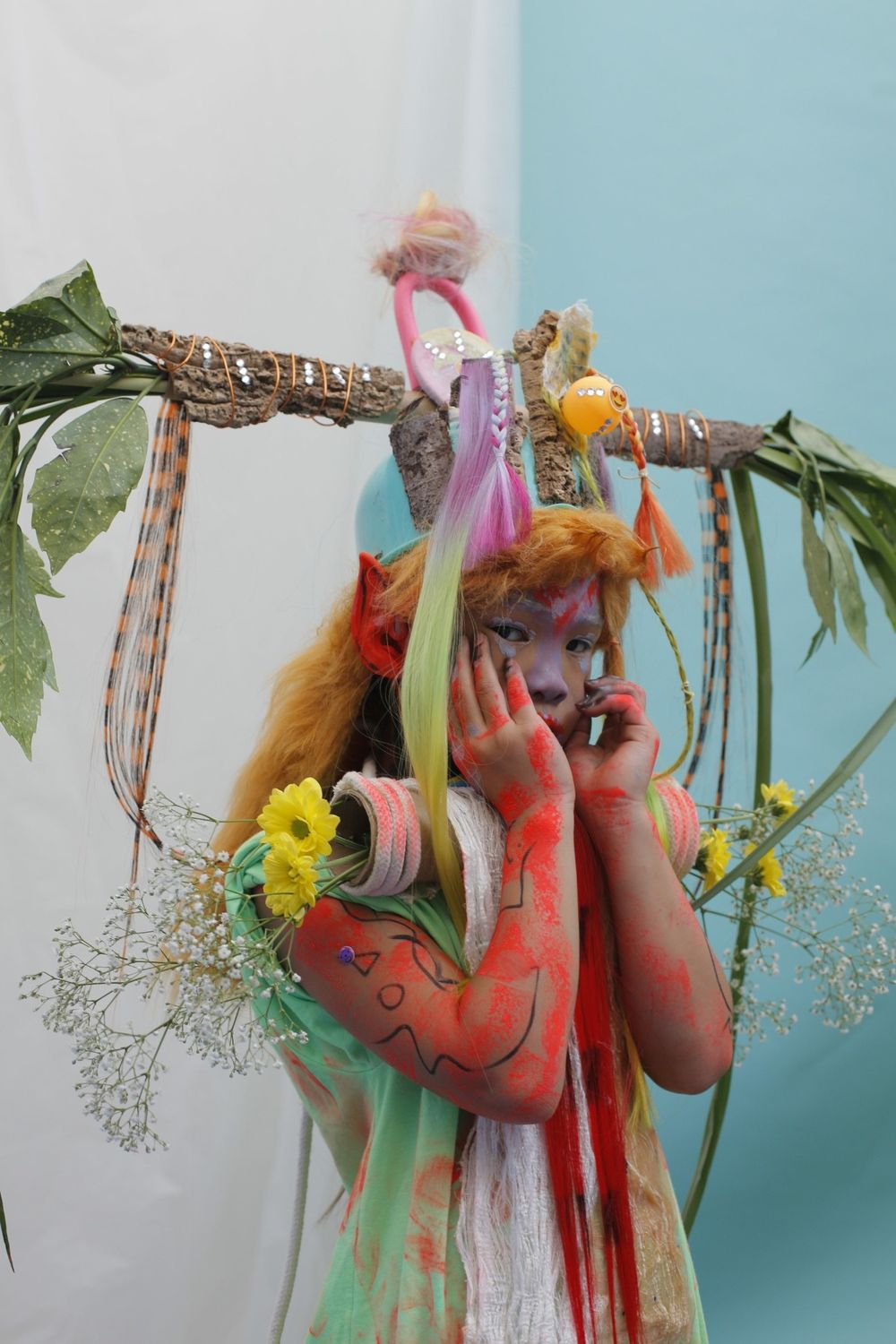 Portret van een figuur in fantasierijke kostuums met bloemen, felgekleurde verf en natuurlijke materialen, gefotografeerd tegen een lichtblauwe achtergrond.