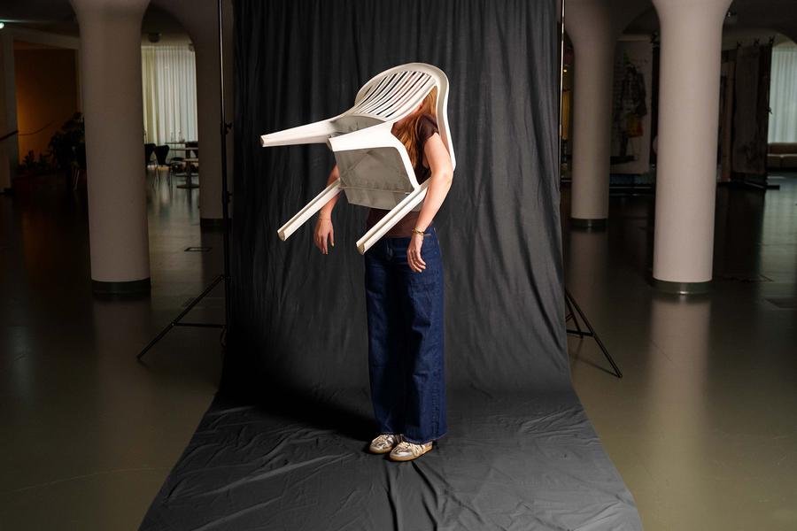 Persoon staat voor een donkere achtergrond terwijl een witte kunststof stoel over het hoofd en bovenlichaam is geplaatst. De foto speelt op een creatieve en surrealistische manier met vorm, object en lichaam in een grote binnenruimte met zuilen.