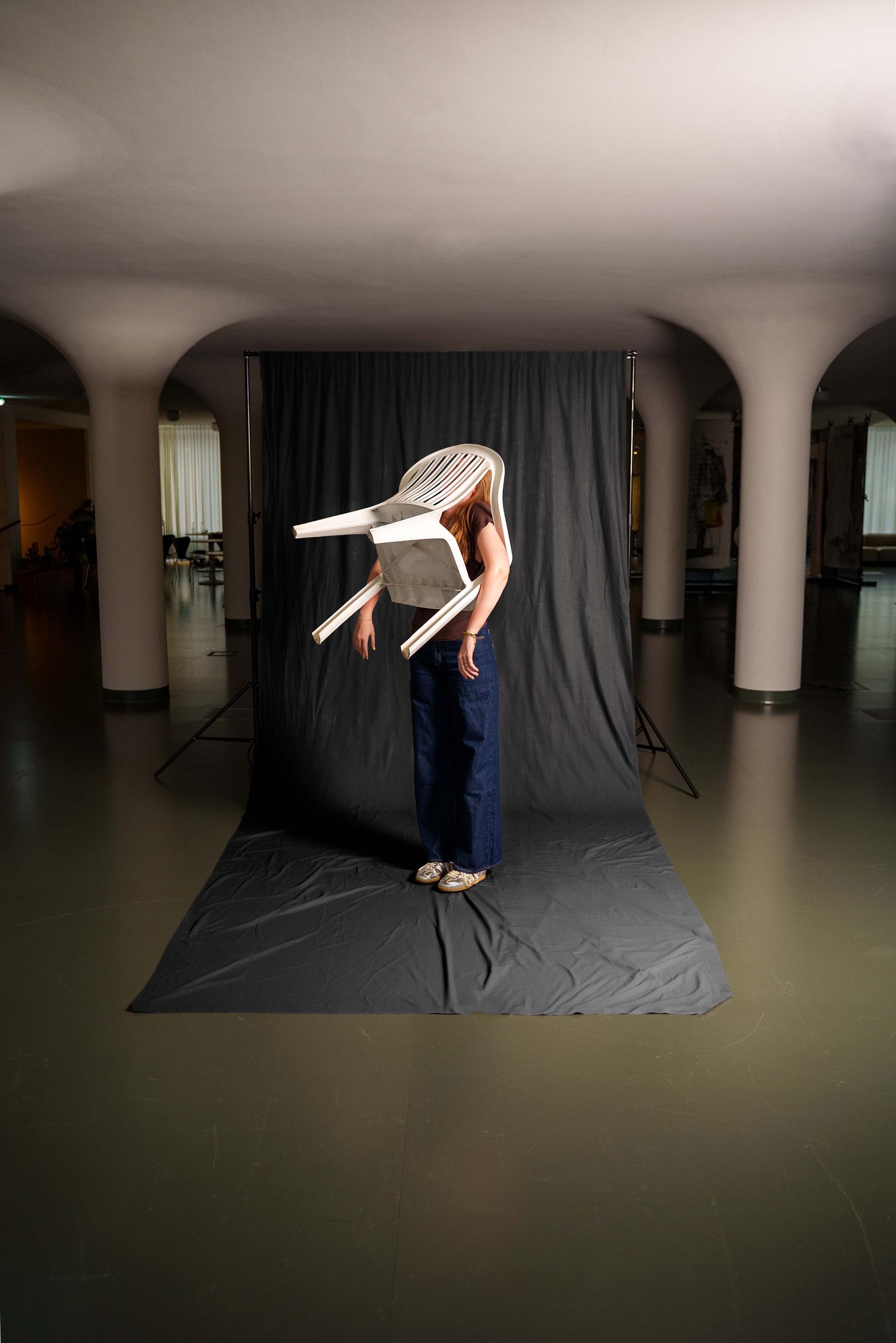 Persoon staat voor een donkere achtergrond terwijl een witte kunststof stoel over het hoofd en bovenlichaam is geplaatst. De foto speelt op een creatieve en surrealistische manier met vorm, object en lichaam in een grote binnenruimte met zuilen.