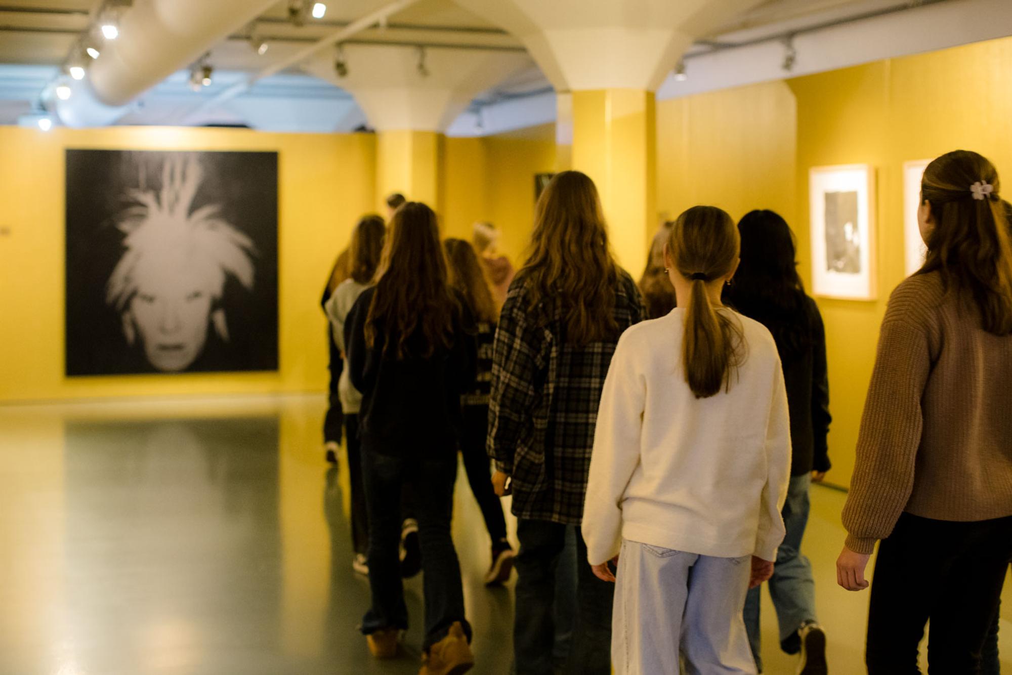 Een groep jongeren loopt door een museumzaal met goudkleurige muren en kunstwerken aan de wand.

