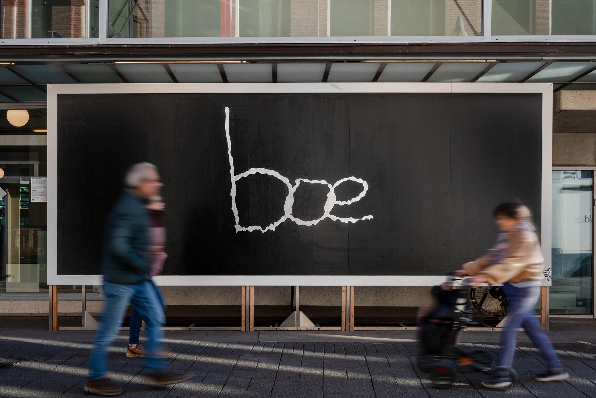 Grote zwarte billboard met het woord “boe” in witte lijnen; op de voorgrond lopen en fietsen voorbijgangers langs.