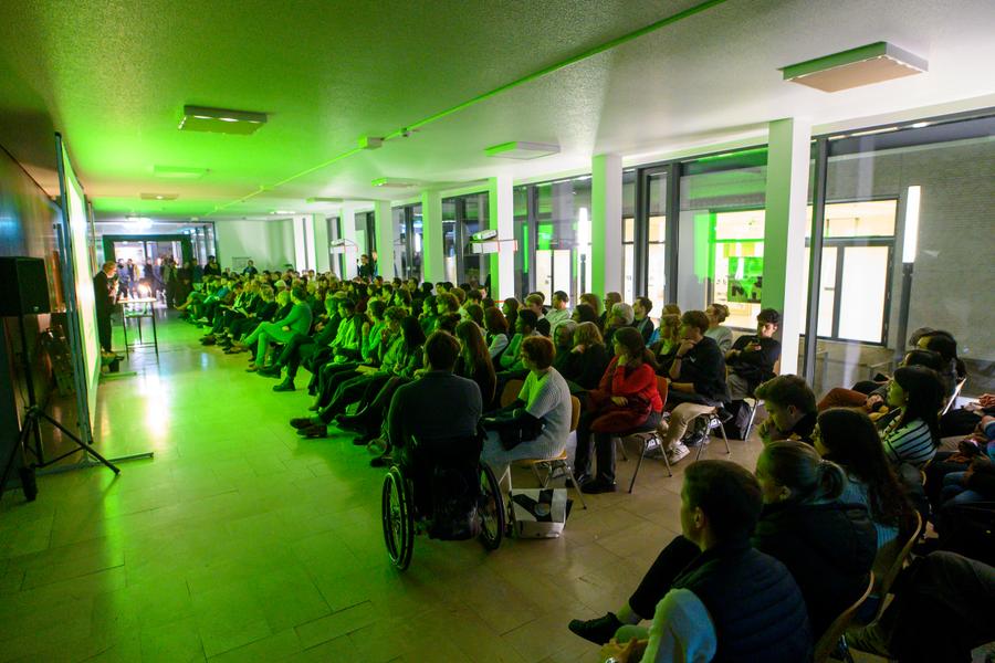 Een volle zaal met publiek dat aandachtig luistert naar een presentatie, onder sfeervol groen licht, in een moderne en open ruimte.