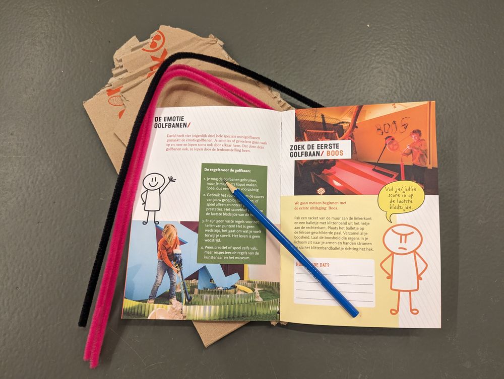 Opengeslagen Museumtas Doe-Boekje met zichtbare creatieve opdrachten, omringd door een potlood en knutselmateriaal zoals papier, folie en een schaar, klaar voor gebruik.
