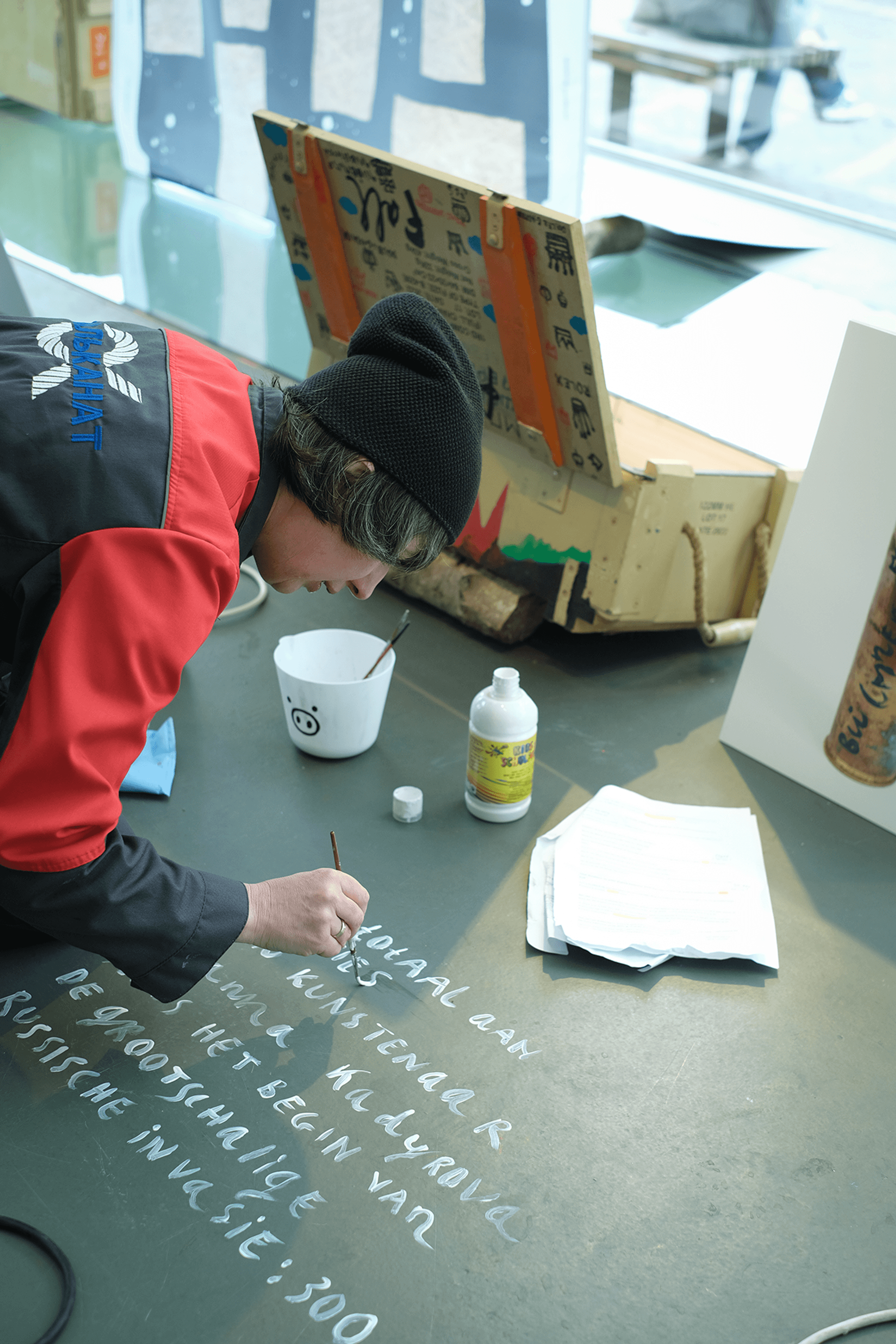 Persoon met rode jas en zwarte muts schildert witte letters op een vloer; naast hen staan een witte emmer met een varkensgezicht, een fles verf en enkele papieren.