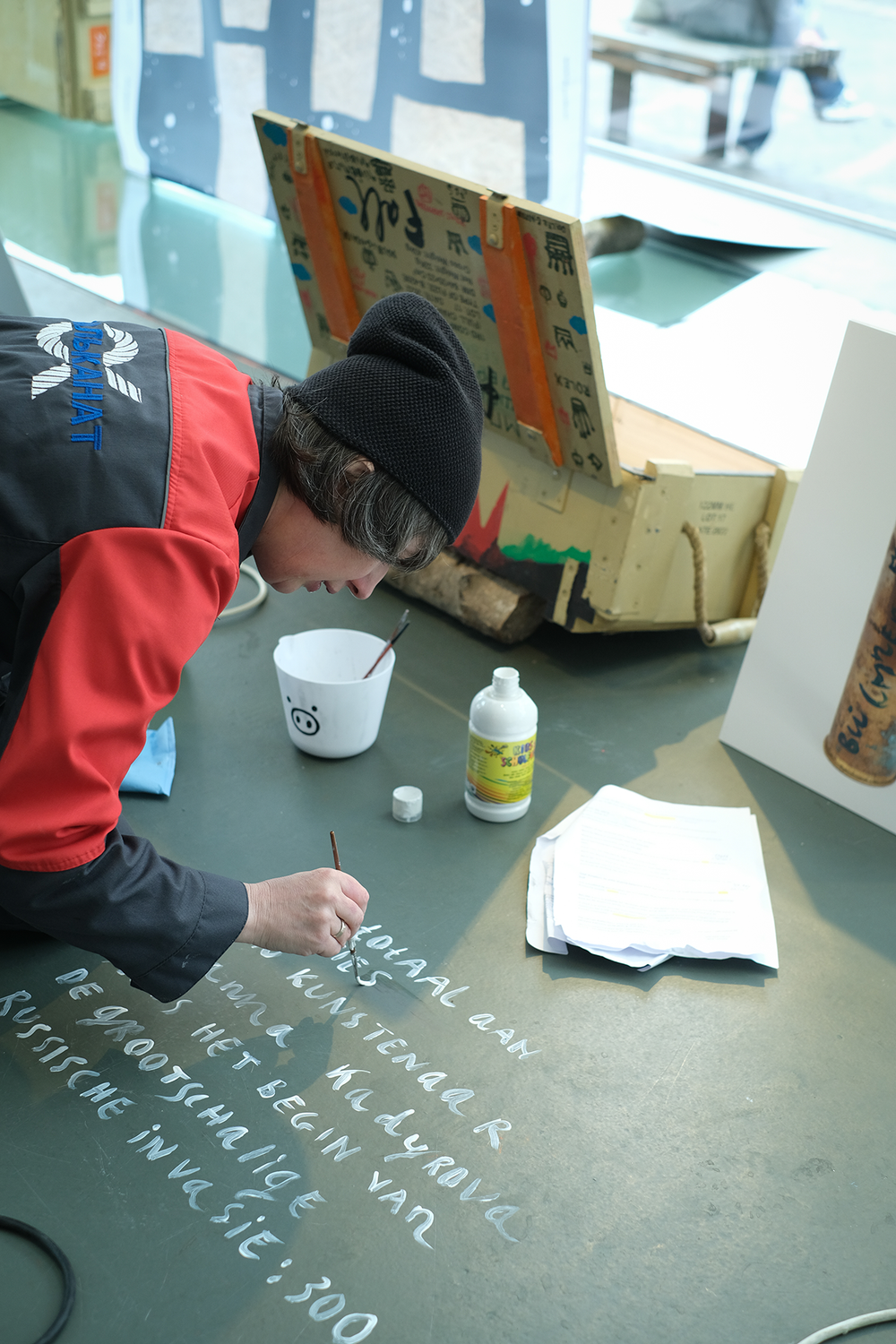 Persoon met rode jas en zwarte muts schildert witte letters op een vloer; naast hen staan een witte emmer met een varkensgezicht, een fles verf en enkele papieren.