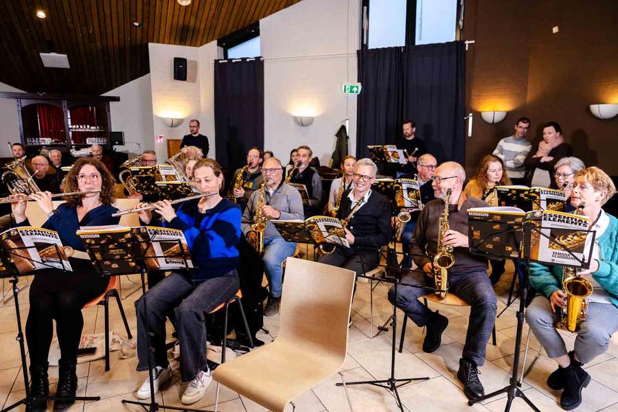 Leden van een harmonieorkest repeteren in een zaal en bespelen onder meer dwarsfluiten, saxofoons en koperinstrumenten. Voor de muzikanten staan lessenaars met bladmuziek opgesteld.