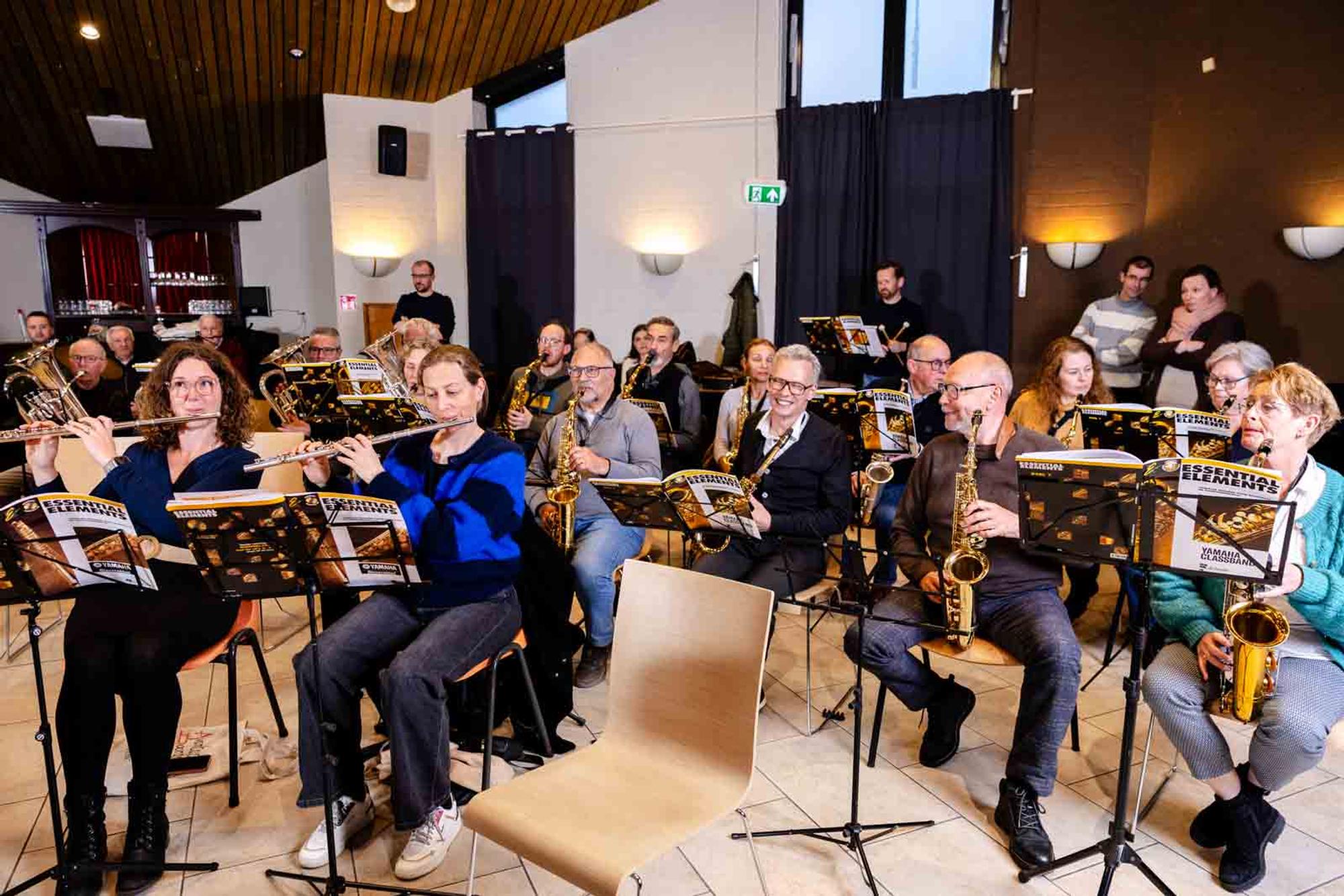 Leden van een harmonieorkest repeteren in een zaal en bespelen onder meer dwarsfluiten, saxofoons en koperinstrumenten. Voor de muzikanten staan lessenaars met bladmuziek opgesteld.
