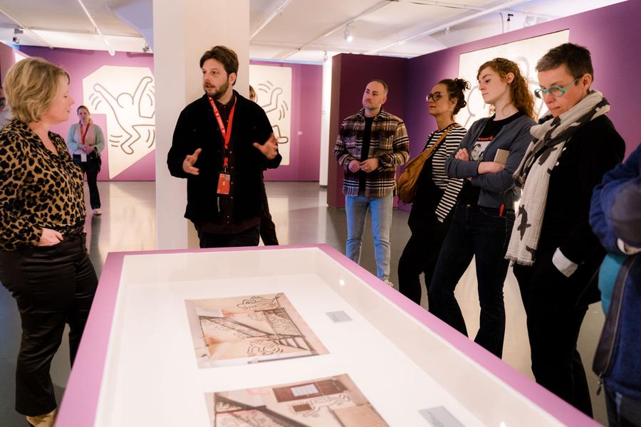 Een groep mensen in een kunstgalerie verzameld rond een vitrinekast met twee kunstwerken. Eén persoon lijkt de kunstwerken aan de groep uit te leggen. Aan de muren van de galerie hangen nog meer kunstwerken.