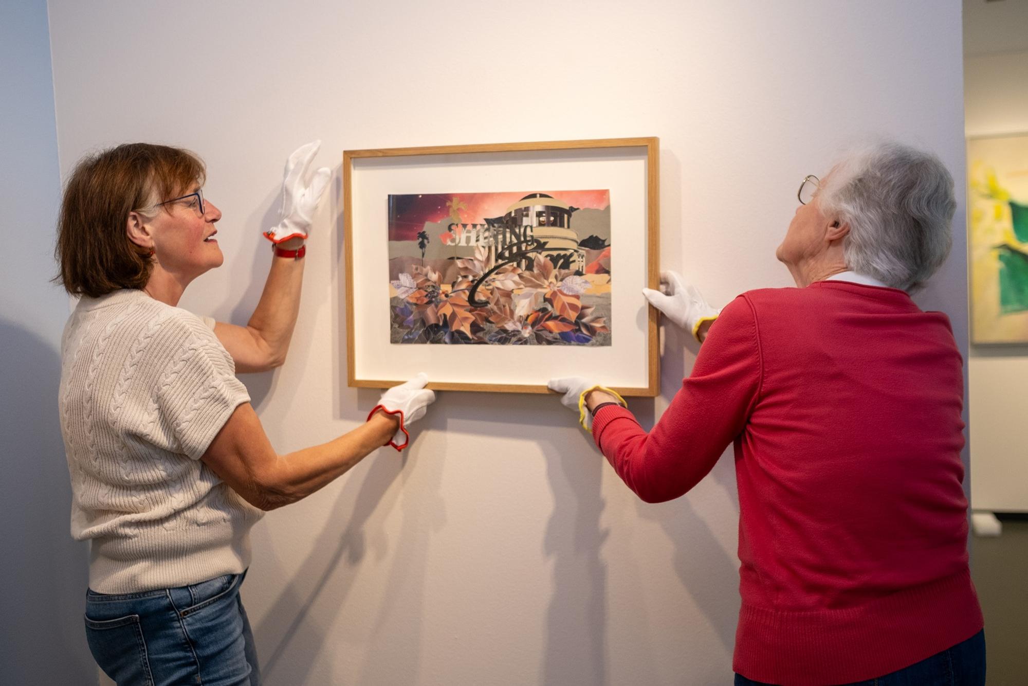 Twee vrouwen hangen een schilderij op tegen een witte muur.