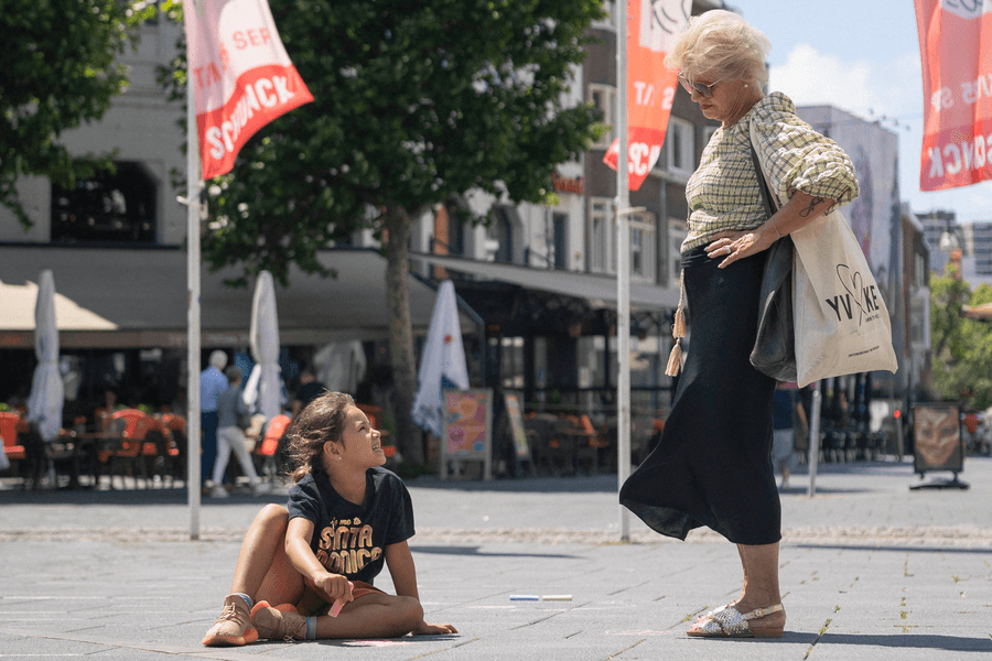 Meisje zit lachend op de grond met stoepkrijt terwijl een oudere vrouw met boodschappentas toekijkt op het Pancratiusplein in Heerlen, met SCHUNCK-vlaggen op de achtergrond.