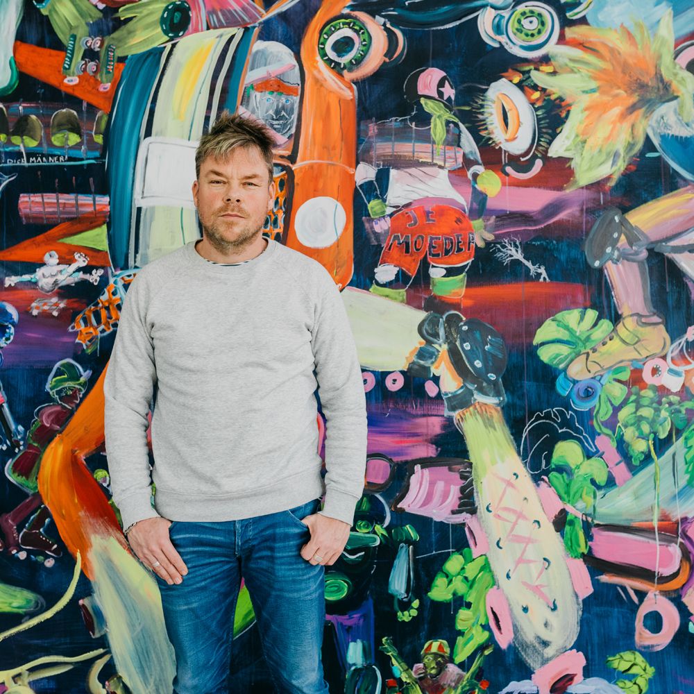 David Bade in grijze trui staat met zijn handen in zijn broekzakken voor een kleurrijk schilderwerk.