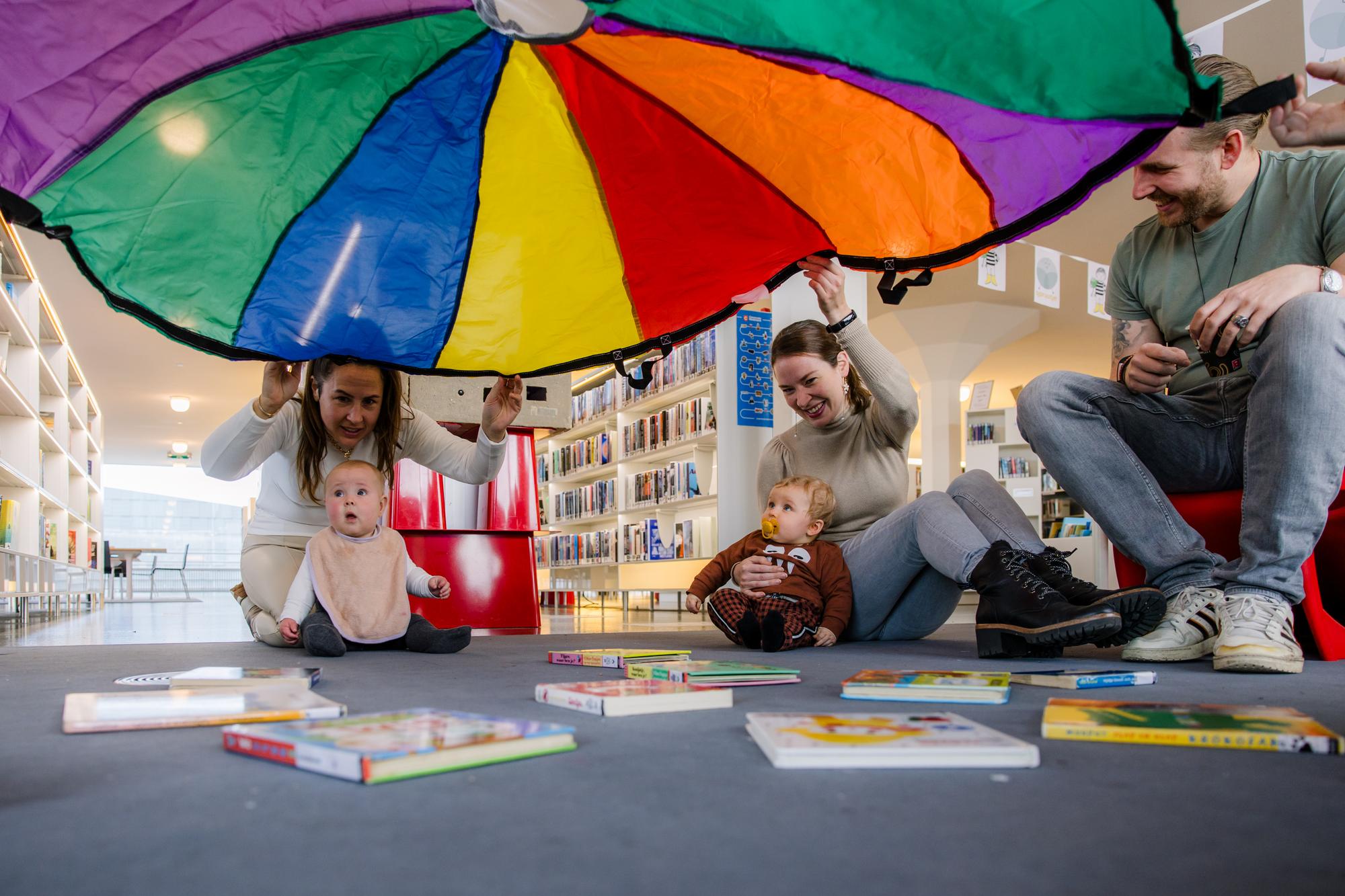 Twee baby’s zitten met hun ouders op de vloer van een bibliotheek onder een kleurrijke parachutedoek, omringd door prentenboeken.