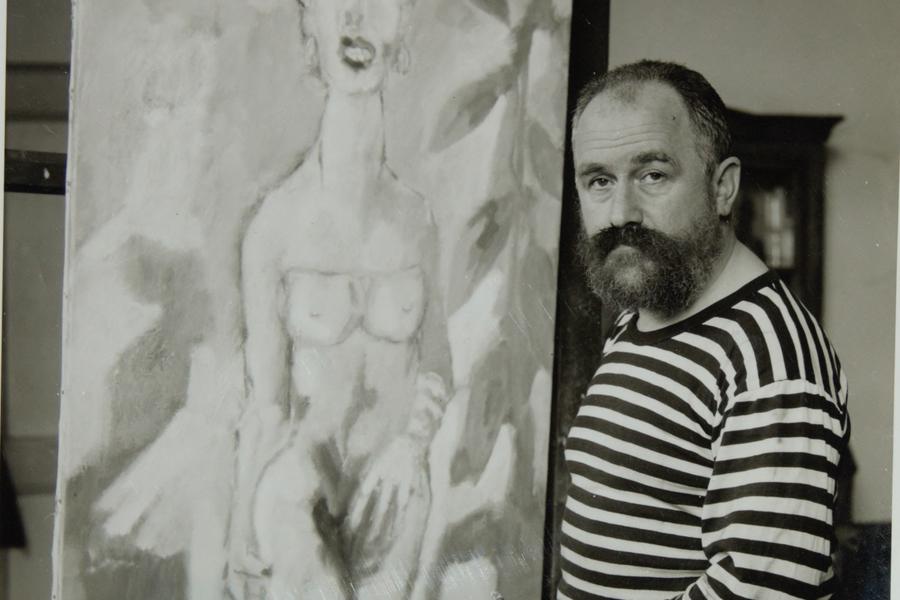 Zwart-witfoto van Aad de Haas, een man met gestreept shirt en baard, in een atelier naast een schilderij van een naakte vrouw op een ezel.