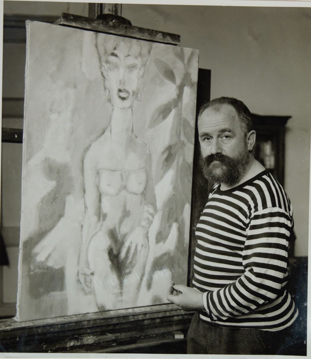 Zwart-witfoto van Aad de Haas, een man met gestreept shirt en baard, in een atelier naast een schilderij van een naakte vrouw op een ezel.
