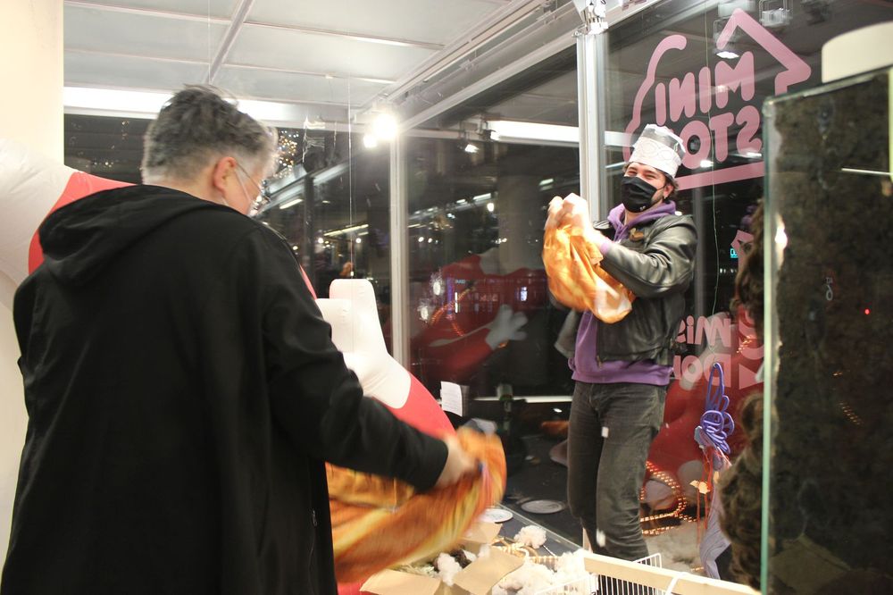 Twee mensen staan in de vitrine van SCHUNCK met mondkapjes op en oranje doeken, lege zakken in de vorm van een baguette, in hun handen. Een van hen draagt een koksmuts. Op de grond liggen spullen. 