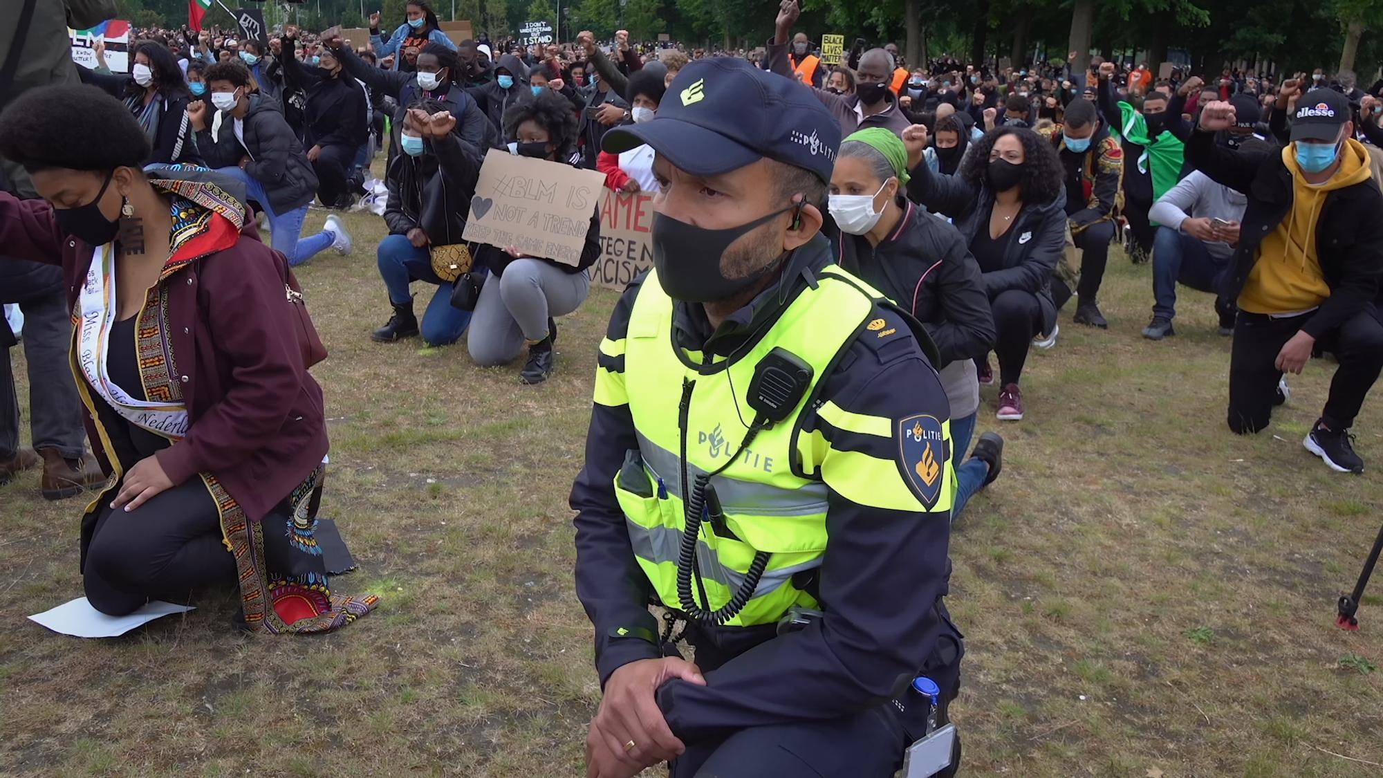 Menigte knielt tijdens demonstratie. Op de voorgrond knielt een politieagent, die een mondkapje draagt.