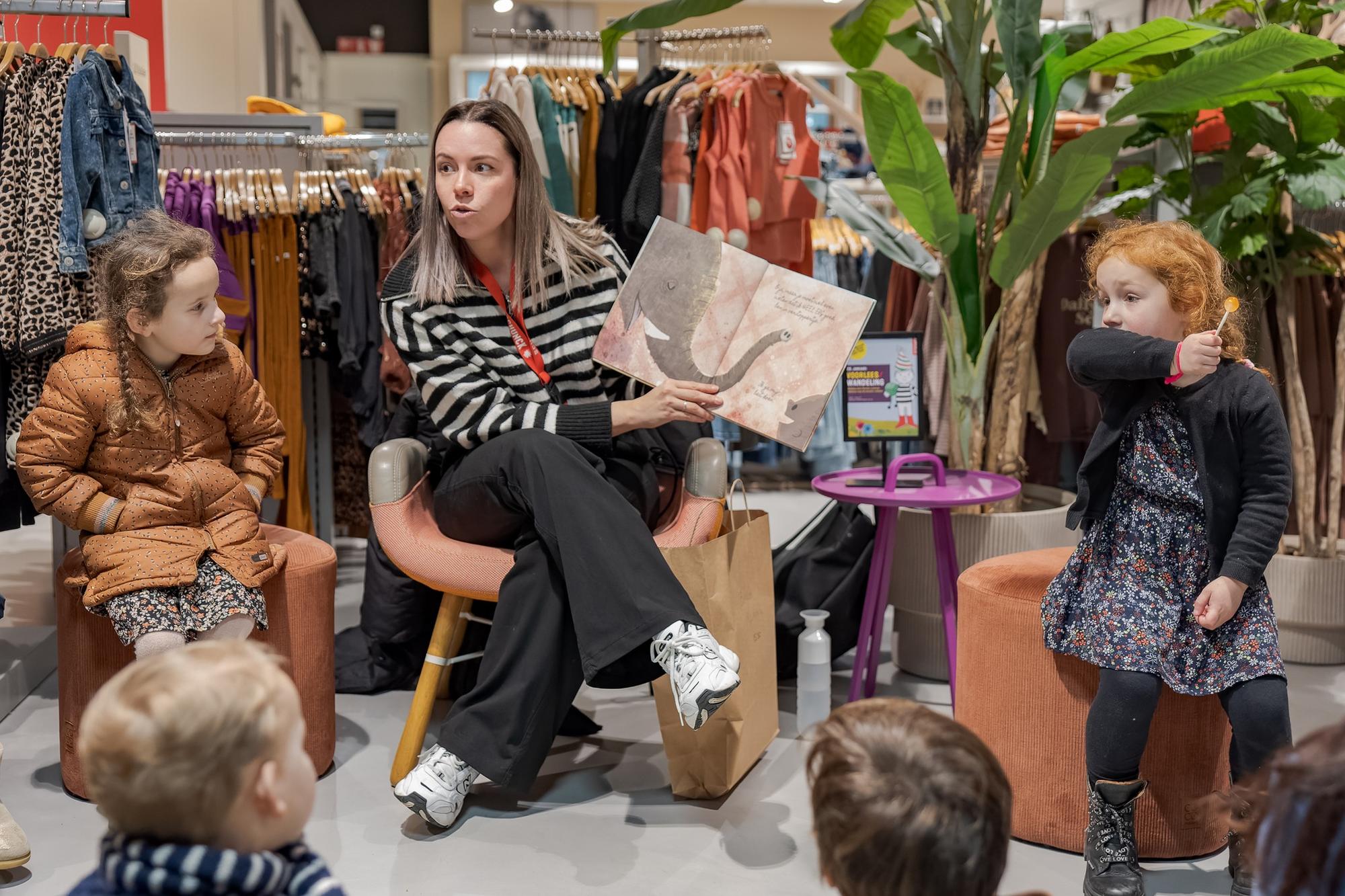 Een vrouw die zittend voorleest uit een boek terwijl ze een afbeelding laat zien en een aantal kinderen die naar haar kijken. 