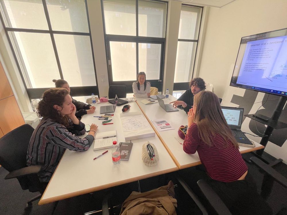 Vijf studenten zitten rond een tafel en bespreken hun ideeën. Op tafel liggen laptops, schetsen en notitieboeken. Rechts is een presentatie op een scherm te zien.