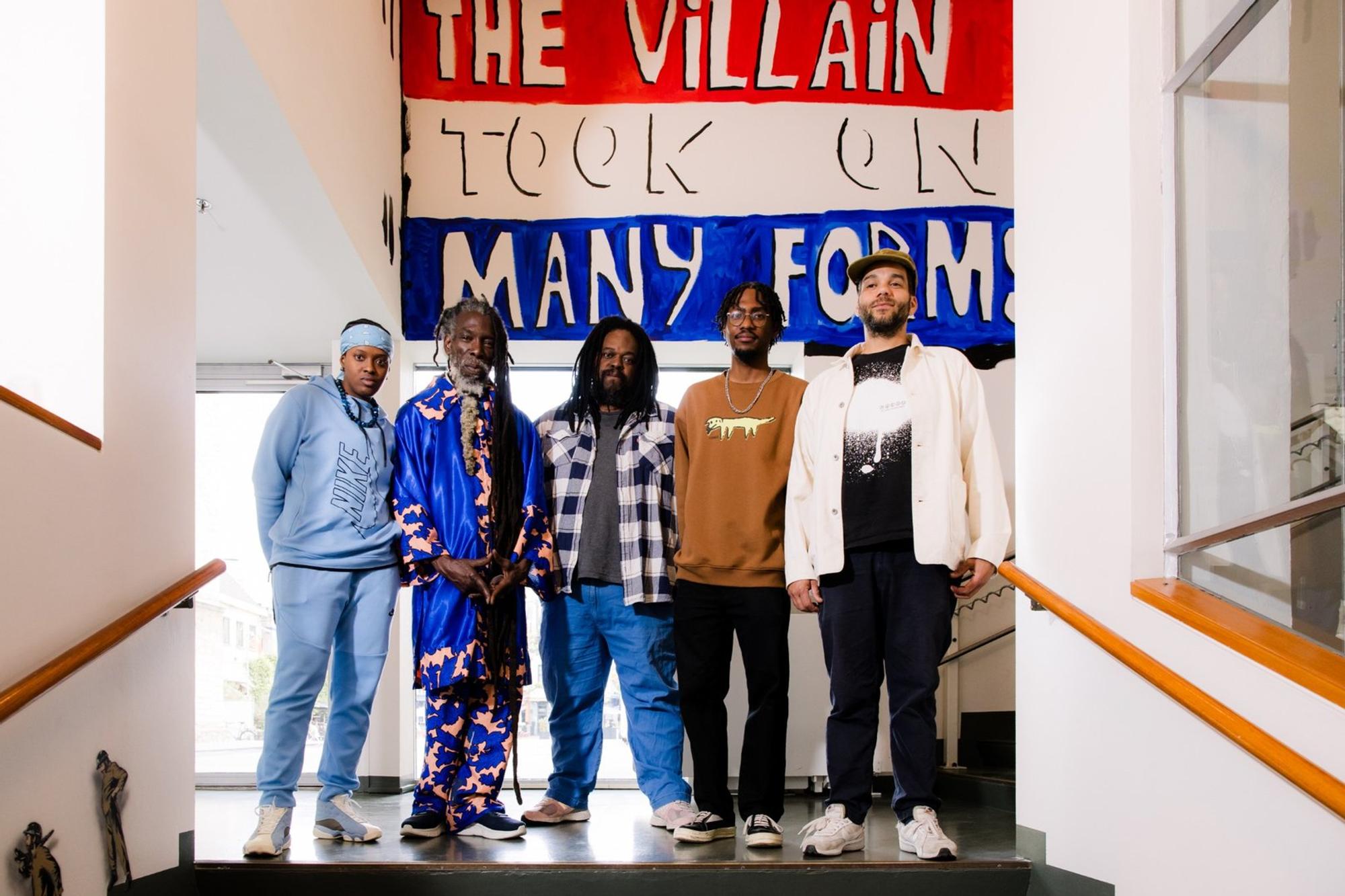 Deelnemende kunstenaars en curator van 'De Politiek van het Dagelijks Leven' poseren voor muurschildering met de tekst 'The villain took on many forms'.