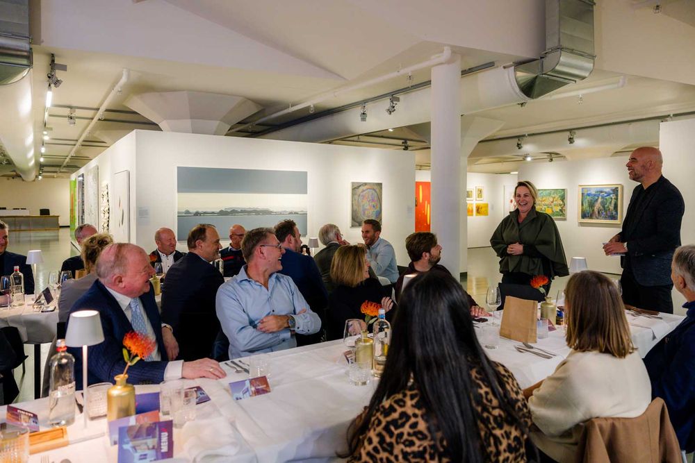 Mensen zitten en praten met elkaar aan een lange tafel in het museum. Ze kijken naar twee personen die staan.