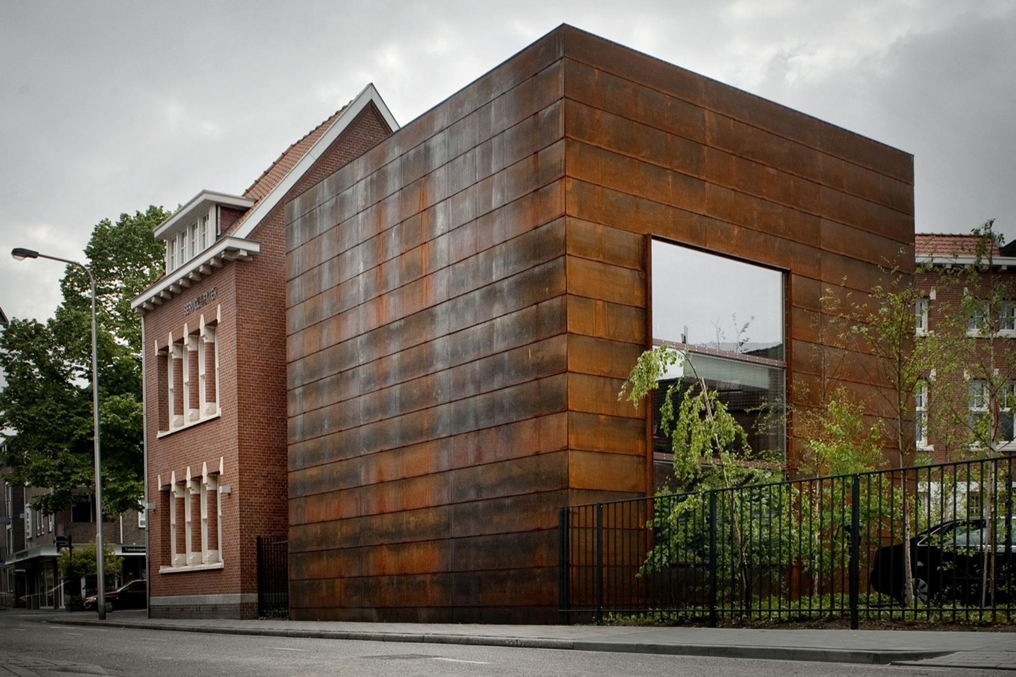 Een modern gebouw met een gevel van roestkleurige cortenstalen panelen, naast een traditioneel bakstenen pand met witte kozijnen en een puntdak. Het moderne deel heeft een groot vierkant raam en staat achter een zwart metalen hek, met bomen en planten zichtbaar in de tuin.