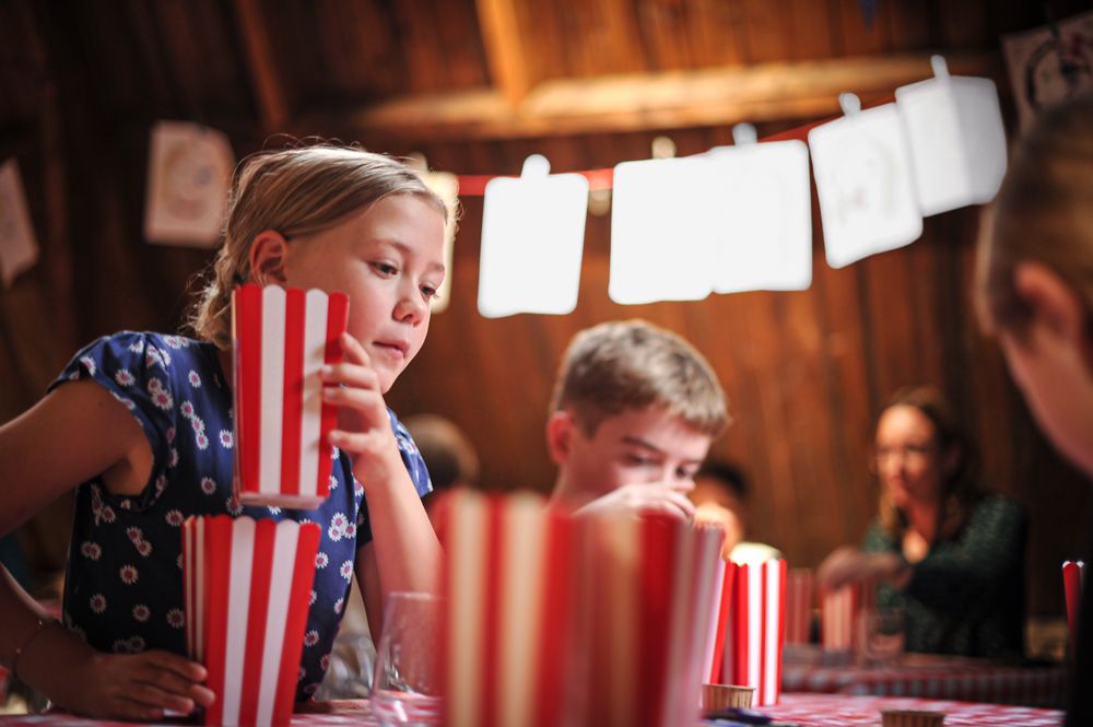Kinderen vullen rood-wit gestreepte popcornbakjes tijdens het kinderboekenfeestje van Bob Popcorn in een warme, houten ruimte. Op de voorgrond stapelt een meisje geconcentreerd de bakjes op tafel terwijl andere kinderen op de achtergrond meedoen aan de activiteit.
