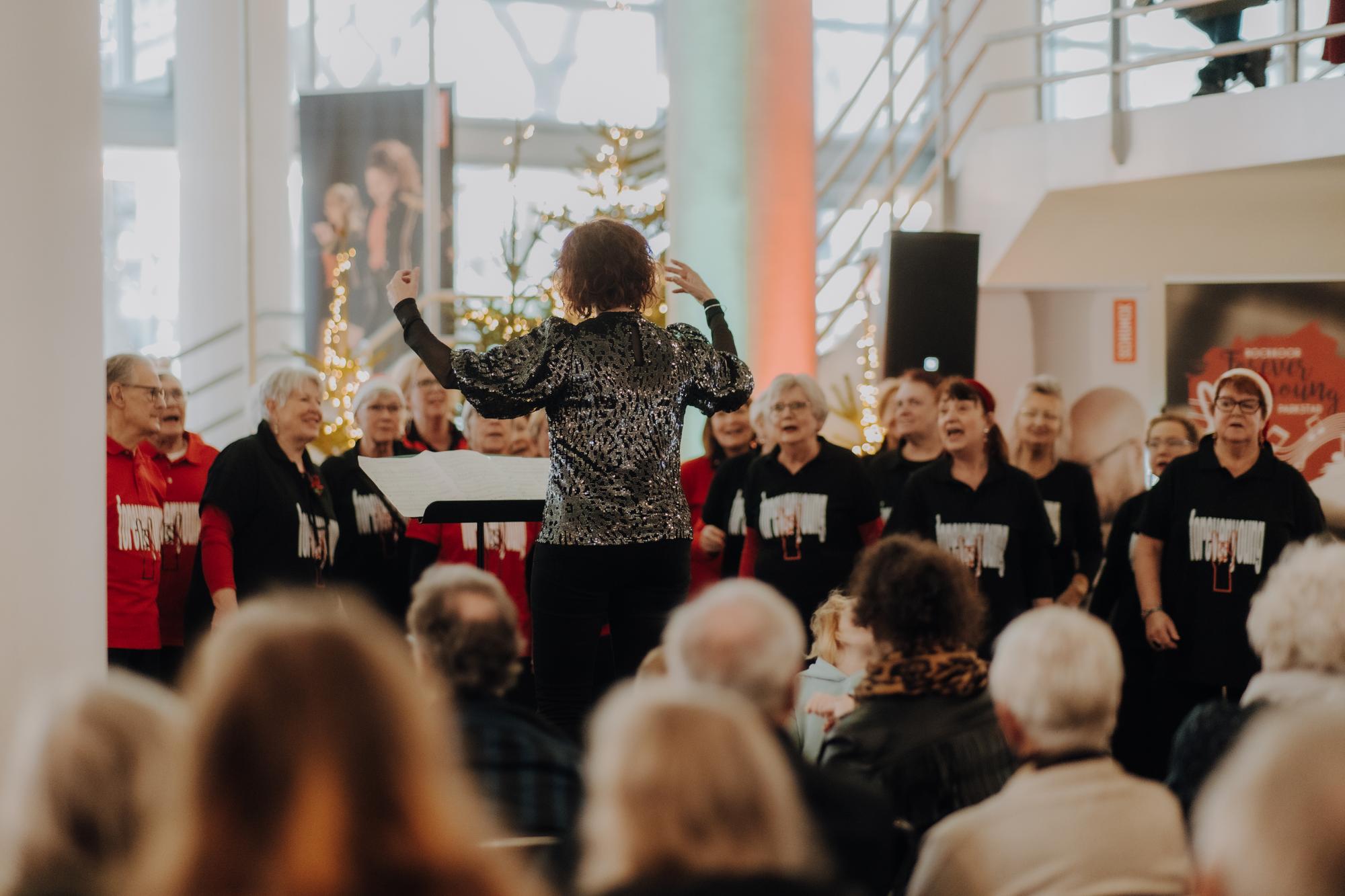 Dirigent staat voor een koor die optreedt tijdens het winterconcert in de hal van SCHUNCK, met onscherpe bezoekers op de voorgrond die toekijken.