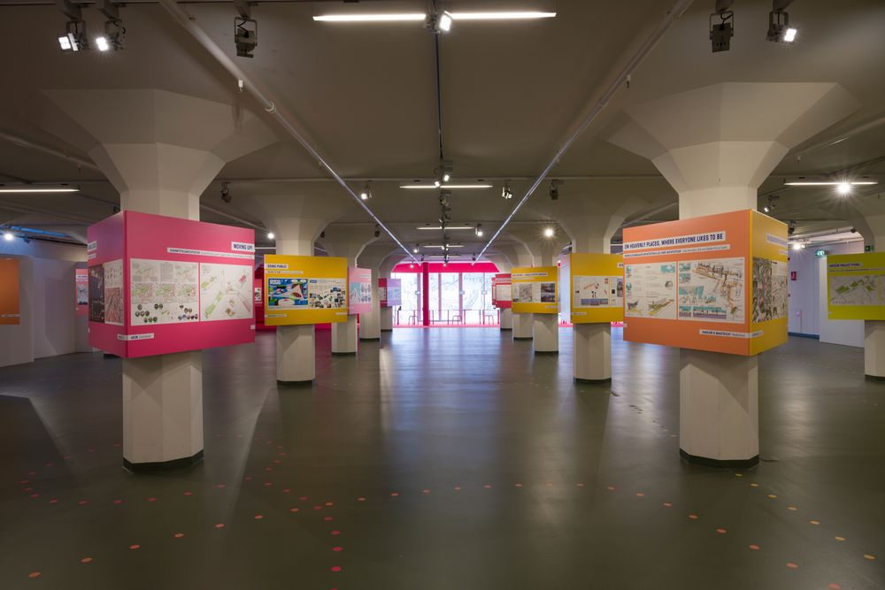  Overzicht van een tentoonstelling met kleurrijke presentatiepanelen op zuilen in een grote zaal; informatieborden tonen kaarten, ontwerpen en teksten.